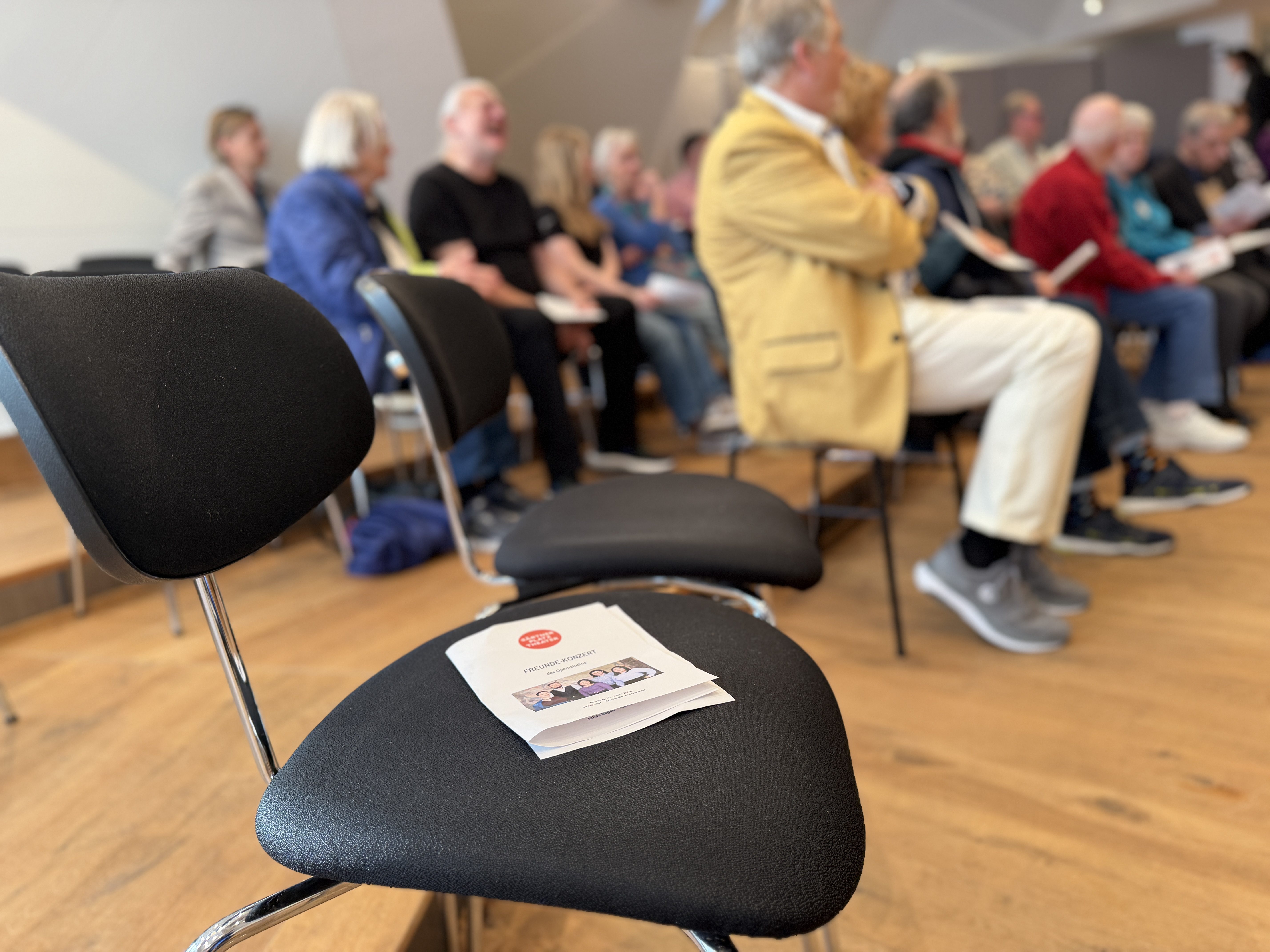 Auf dem Stuhl liegt ein Programmzettel, dahinter sind unscharf Menschen im Publikum